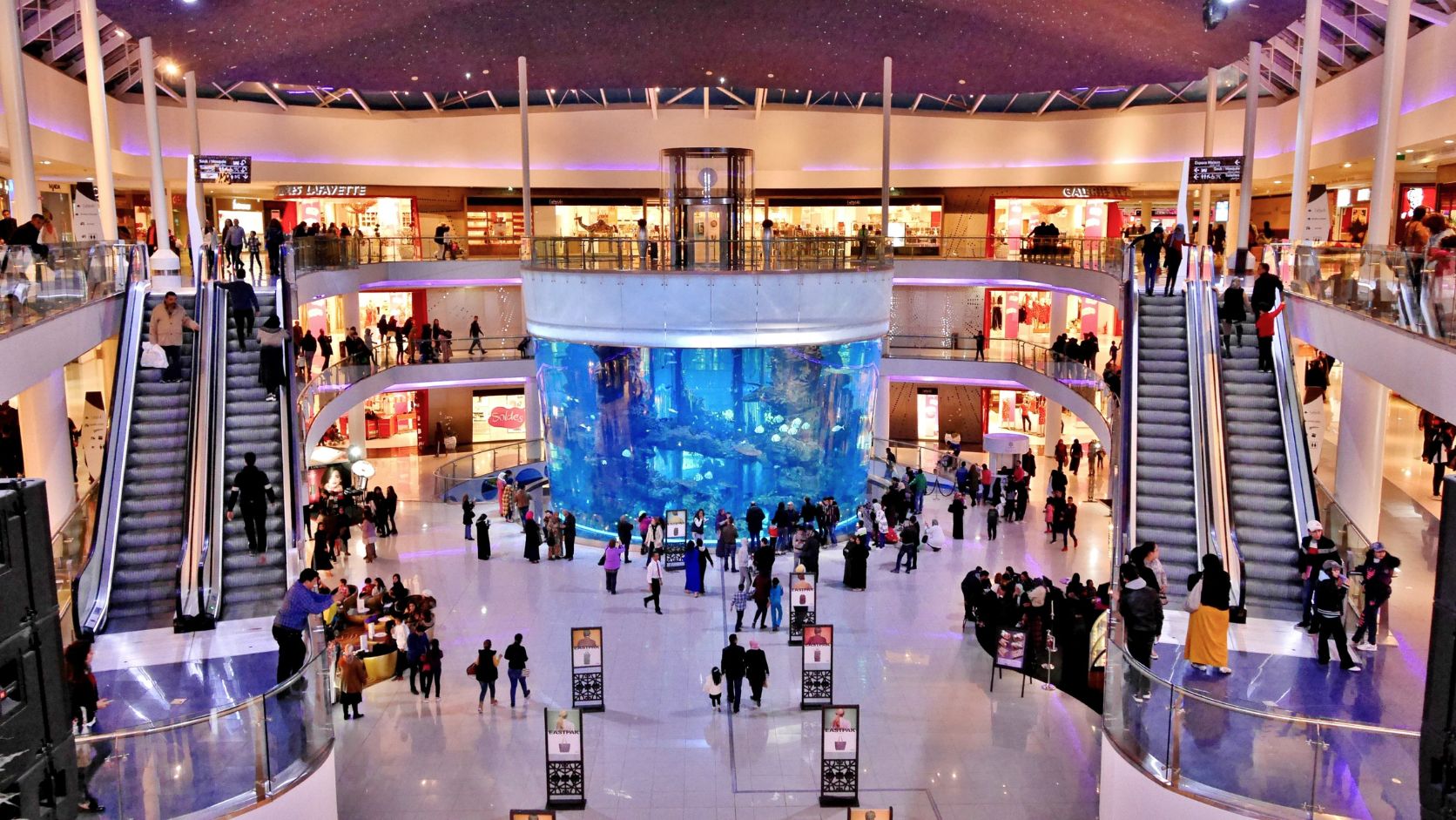 Interior de un centro comercial de varios niveles con escaleras eléctricas, iluminación moderna y tiendas alrededor de un espacio central.